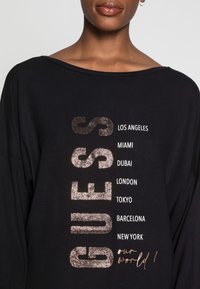 Schwarzes Langarmshirt mit weitem Ausschnitt. Auf der Vorderseite befindet sich metallischer Glitzertext mit Städtenamen und dem Satz "unsere Welt!" in Kursivschrift.