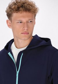 Giacca a cappuccio navy con zip frontale che presenta accenti azzurri. Texture morbida; design aderente, indossata sopra una camicia bianca.