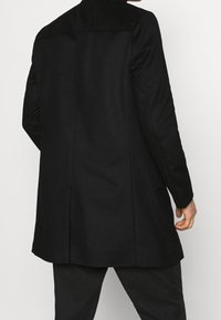 Cappotto lungo nero con una vestibilità sartoriale, texture liscia e un'apertura sul retro. Presenta un delicato dettaglio di cucitura lungo le spalle.