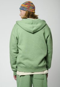 Haut à capuche vert au style décontracté, doté d'une grande capuche et d'un ourlet à côtes. Porté sur une chemise couleur crème, assortie à un pantalon vert.