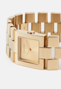 Reloj de metal dorado con caja cuadrada, esfera dorada plana y marcadores de hora minimalistas. Presenta una pulsera rectangular unida y grabado de logotipo.