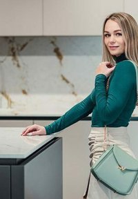 Zelený žebrovaný top s dlouhým rukávem, smetanová zavinovací sukně, držící mátově zelenou kabelku se zlatými detaily, na pozadí mramorové kuchyně.