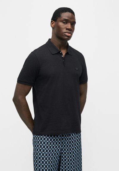 Άνδρας που φοράει μαύρο πόλο με κοντά μανίκια και μπλε-μαύρο παντελόνι με σχέδια, στέκεται με τα χέρια πίσω από την πλάτη μπροστά σε λευκό φόντο.