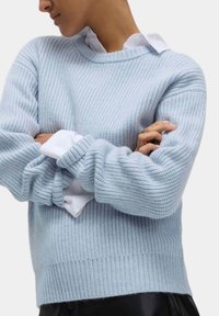 Jasnoniebieski sweter z dzianiny o prążkowanej teksturze, okrągłym dekolcie i długich rękawach, noszony na białej koszuli z kołnierzykiem, w wersji skróconej.