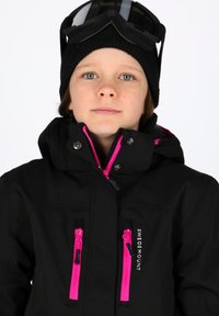 Giacca da sci nera con zip e dettagli rosa, colletto alto e logo del marchio. Abbinata a un berretto di lana nero e occhiali da sci.