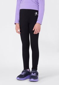Leggings neri realizzati in tessuto elastico, con un piccolo logo bianco sulla vita. Abbinati a calzature viola.