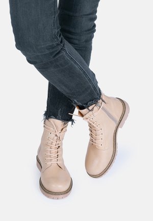 Beige nahkajalkineet, joissa on nauhakiinnitys, kumipohjat ja ommellut yksityiskohdat. Yhdistetty tummiin farkkuihin, joissa on revitty lahje.