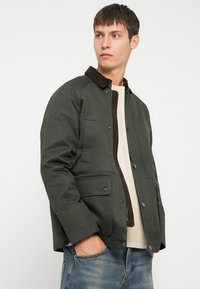 Olijfgroene jas met een bruine corduroy kraag, met knoopsluitingen en twee voorzakken, gedragen over een beige top en spijkerbroeken.