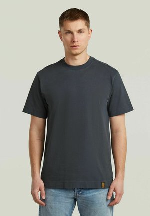 Giovane uomo con capelli corti che indossa una t-shirt a maniche corte grigio scuro semplice e jeans azzurri, in piedi davanti a uno sfondo semplice.