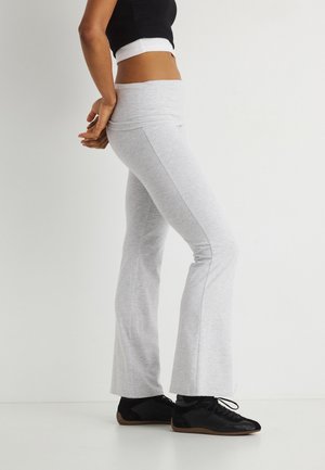 Persoană purtând pantaloni evazați gri deschis, top scurt negru și pantofi negri cu șireturi, stând lateral pe un fundal alb.