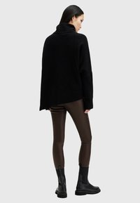 Kobieta ubrana w czarny, oversize'owy sweter z wysokim kołnierzem, ciemnobrązowe dopasowane skórzane spodnie i czarne botki na kostkę, stojąca na jednolitym tle.