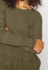 Olivgrüner Pullover mit langen Ärmeln und einer gerippten Struktur, ausgestattet mit einem Rundhalsausschnitt und einem lockeren Schnitt, dargestellt mit verschränkten Armen.