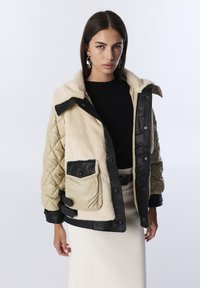 Femme portant une veste matelassée beige et noire avec un devant en shearling et une jupe crème, posant devant un fond blanc uni.
