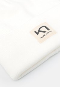 Berretto di maglia bianco con risvolto, dotato di un'etichetta quadrata beige con testo nero "K1 KARI TRAA". Texture liscia e design minimale.