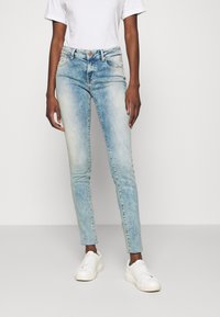 Jeans de denim azul claro con un acabado desgastado, diseño de alta cintura, estilo de cinco bolsillos y un corte ajustado, combinados con zapatillas blancas.