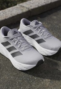 Ζευγάρι γκρι παπούτσια τρεξίματος Adidas με μαύρες ρίγες και λευκές σόλες τοποθετημένα σε άσφαλτο κοντά σε πέτρινο κράσπεδο.