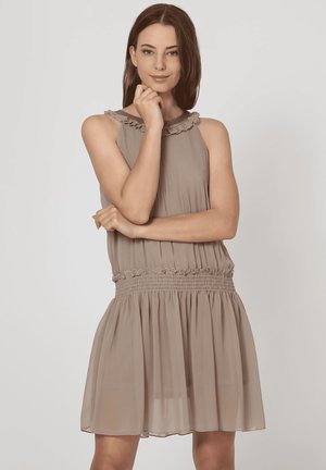 Joven mujer con pelo castaño lacio vistiendo un vestido de gasa taupe sin mangas, posando con una mano tocándose la barbilla y la otra cruzada.