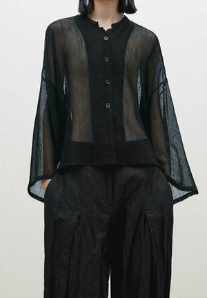 Femme portant un chemisier noir ample et transparent avec des manches larges et un pantalon noir ample assorti, mains dans les poches.