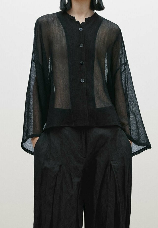 Femme portant un chemisier noir ample et transparent avec des manches larges et un pantalon noir ample assorti, mains dans les poches.