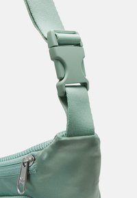 Zelený nylonový popruh s plastovou sponou, který má stříbrný kovový jezdec na zip pro bezpečné uzavření a texturovaný povrch látky.