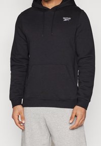 Sweat à capuche noir en tissu doux, avec une poche kangourou à l'avant, des cordons de serrage et un petit logo Reebok blanc sur la poitrine.