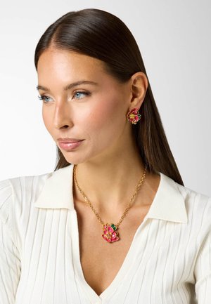 Frau mit langen braunen Haaren, die ein weißes, geripptes Oberteil, eine goldene Halskette und passende florale Ohrringe mit rosa und grünen Akzenten trägt.