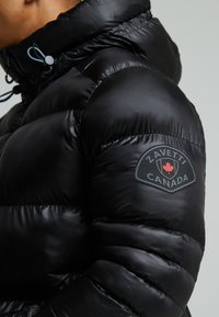 Čierna puffová bunda so lesklou textúrou má vysoký golier a nastaviteľné šnúrky. Vyšité logo Zavetti Canada na rukáve.