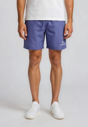 Mężczyzna ubrany w niebieskie spodenki Sergio Tacchini, białe tenisówki i białą koszulkę, stojący na tle jednolitego szarego tła.