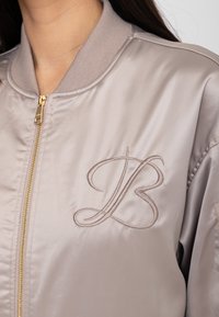 Close-up van een persoon die een licht beige satijnen bomberjack draagt met een gouden rits en geborduurde, in elkaar verstrengelde letters "LB" op de borst.