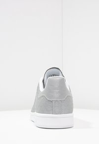 Zapatilla gris con parte superior texturizada, acentos blancos y suela de goma plana. Cuenta con un collar acolchado y un logo en relieve en el talón.