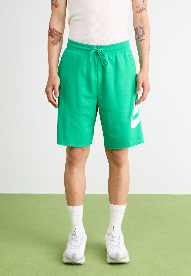 Shorts de sport verts en tissu doux, avec une taille élastique et un logo Nike blanc sur la jambe gauche. Portés avec des baskets blanches.