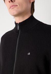 Sweater noir à fermeture éclair avec un col côtelé et des détails de fermeture éclair en argent. Présente un petit logo "ck" blanc sur la poitrine. Texture de tissu douce.