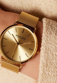 Gold watch with a mesh strap, featuring a round face, textured golden dial, and slim hour markers. Brand name displayed prominently.