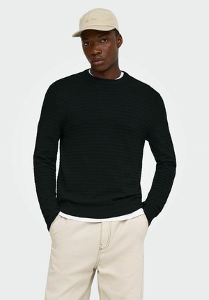 Homme portant une casquette de baseball beige, un pull texturé noir par-dessus une chemise blanche, et un pantalon beige, les mains dans les poches, debout devant un fond clair.