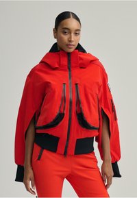 Piros szigetelt dzseki nagy fekete cipzáras zsebekkel, túlméretezett kapucnival és bordázott mandzsettákkal. Simaságú anyag, sportos kialakítás.