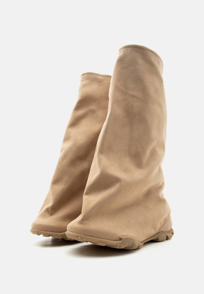 Μαλακά δερμάτινα μποτάκια μέσης γάμπας σε χρώμα ταμπά με στρογγυλεμένες μύτες και υφασμένα λαστιχένια πέλματα σε ανοιχτό φόντο.