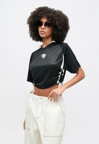 Μαύρο κοντό τοπ με υφασμένο σχέδιο, διαθέτει λευκό γιακά και πλευρικούς δεσμούς. Συνδυάζεται με κρεμ φαρδιά παντελόνια με εμφανείς λεπτομέρειες ραφής.