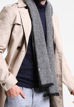 Homme portant un trench-coat beige, une écharpe à carreaux noir et blanc, un col roulé bleu marine et un pantalon gris clair, avec une main dans la poche.