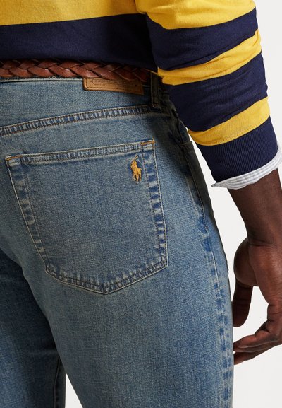 Lyseblå denimjeans med klassisk passform, med en liten gul brodert logo på baklommen og et brunt flettet lærbelte.