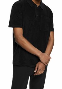 Personne portant un polo noir à manches courtes avec une poche sur la poitrine, les mains jointes devant, sur fond blanc.