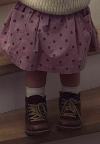 Barn iført en pink prikkeret nederdel, hvide sokker og brun snørebåndsstøvler står på trætrapper.