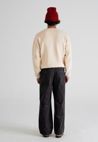 O persoană stă cu spatele, purtând o căciulă roșie din tricot, un pulover crem, pantaloni negri lejeri și pantofi maro, pe un fundal deschis.
