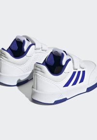 Zapatillas deportivas blancas con acentos azules, que cuentan con correas de Velcro, forro de malla texturizada y detalles perforados en el talón.