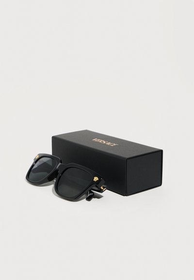 Μαύρα υπερμεγέθη τετράγωνα γυαλιά ηλίου με χρυσά λογότυπα Medusa στα πλάγια, δίπλα σε μαύρο κουτί με το λογότυπο Versace, σε λευκό φόντο.