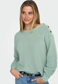 Jeune femme portant un pull en maille vert clair avec des détails de boutons sur l'épaule et un jean bleu, les mains dans les poches, souriant et regardant de côté.