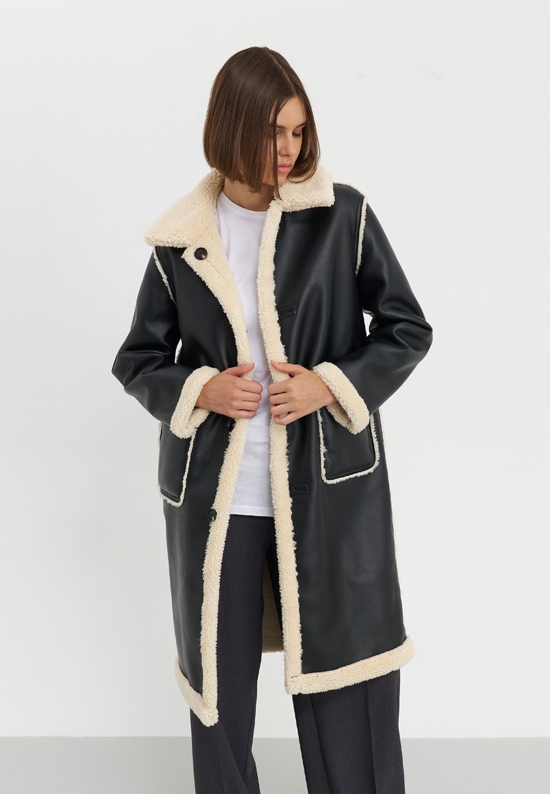 Cappotto in ecopelle nera con fodera in shearling crema, grande colletto, tasche frontali e bordi a contrasto, dotato di chiusura a bottoni.