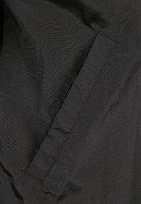 Tejido negro con una textura suave, que presenta una costura de bolsillo horizontal en el lado izquierdo. El material parece ligero y mate.