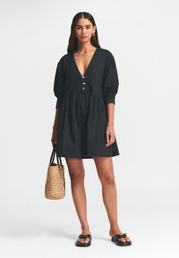 Schwarzes Kleid mit tiefem V-Ausschnitt, Knopfleiste und Puffärmeln. Getragen mit einer geflochtenen beigen Tasche und flachen schwarzen Sandalen. Glatte Textur.