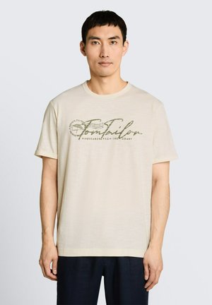 Man draagt beige T-shirt met donkere cursieve "Tom Tailor" tekst en kleine afbeelding, staand tegen een effen lichte achtergrond.