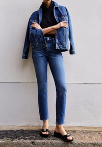 Person wearing blue denim jacket and jeans, black top, and black heeled sandals, standing against a two-tone wall.